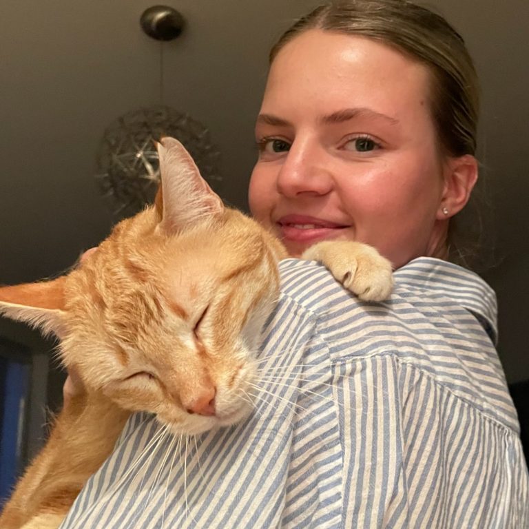 Ein hübsches, lächelndes Mädchen hält eine schmusende, orangefarbene Katze auf der Schulter.